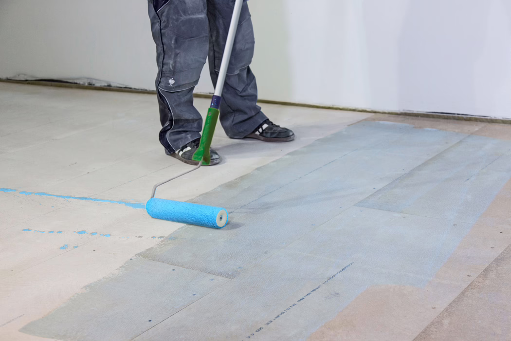 UZIN_Application Image_PE360 Plus_Renoplan System_2020-10 A worker's lower legs and feet are visible while applying a light blue primer or paint to a wooden subfloor with a long-handled paint roller. The person wears dark work trousers and black sneakers; the roller leaves a wet blue coating on the floorboards in an indoor room with plain white walls. The image focuses on the floor preparation process, the tools and paint splatters.