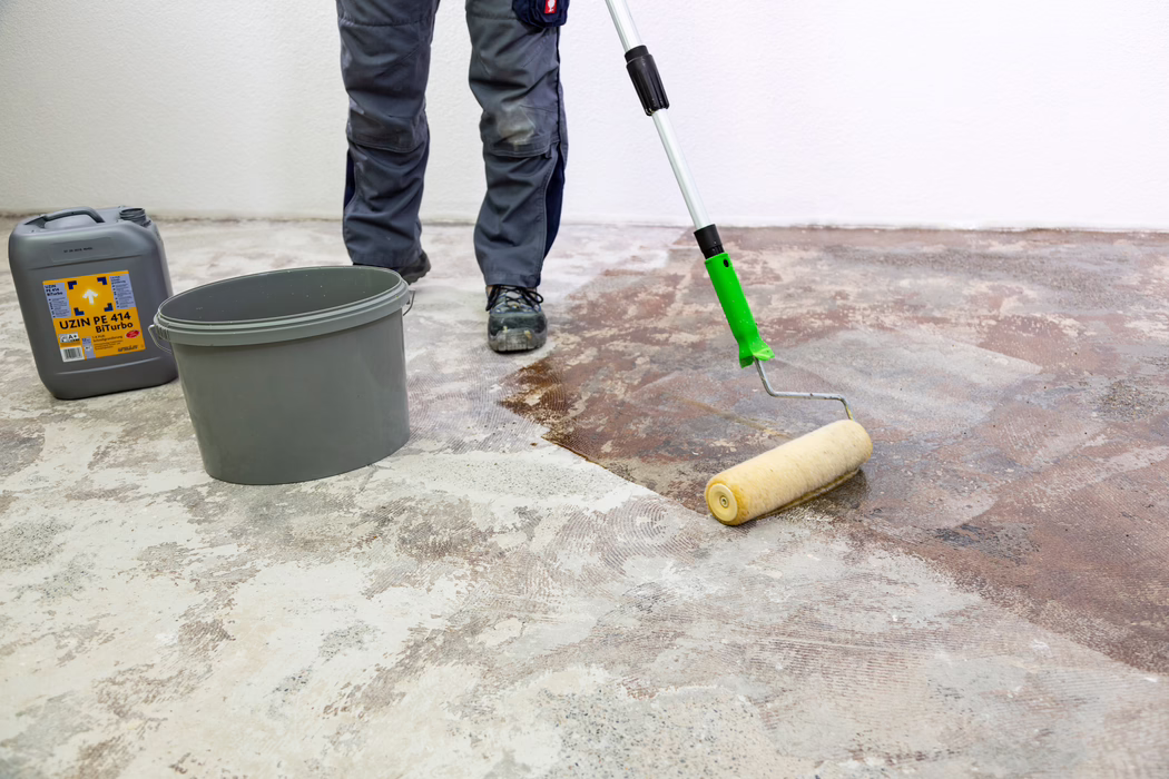 A worker's lower legs and feet are visible as they apply a brown primer or adhesive to a bare concrete floor with a long-handled paint roller. A gray plastic bucket and a gray canister labeled "UZIN PE 414 Biturbo" stand nearby against a white wall. The worker wears gray work trousers and black safety shoes; the floor shows patches of old adhesive and ground concrete while the roller spreads the liquid evenly to prepare the subfloor.