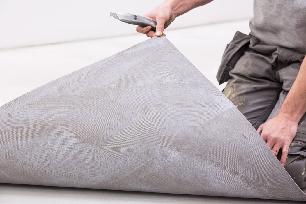A worker kneels and lifts a large sheet of vinyl or linoleum flooring, exposing the textured adhesive trowel ridges on its underside. Only the person's hands, forearms and part of the torso are visible; one hand holds a utility knife while the other supports the rolled edge of the material. The worker wears gray work trousers with paint stains on the knees. The flooring sheet and underlying surface are light gray with circular notched adhesive patterns and small dirt marks, and the image focuses on the close-up detail of hands and flooring rather than the person's face.