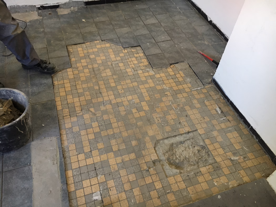 UZIN_Application_Image_UZIN NC 110_0445_ 2018-03 A partially removed tiled floor during renovation: the foreground shows small beige and gray mosaic tiles with worn and missing sections; larger dark rectangular tiles around the edges have been lifted, revealing adhesive and the subfloor. A worker's legs and black boots are visible at left next to a bucket of removed tiles; a red pry bar leans against the right wall. White painted walls with black skirting, dust and debris scattered across the floor. Photo taken from a low angle looking down.