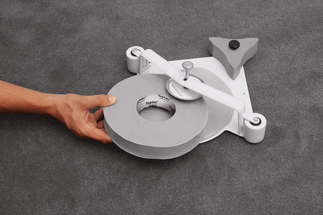 Close-up of a left hand holding a large gray roll of adhesive tape positioned on a low-profile tape dispenser placed on a gray textured fabric surface. The dispenser features a white metal arm with a central spindle and a round cutting wheel, two small white rollers at the base and a triangular gray stabilizer with a black rubber foot; the cardboard core of the tape shows the printed brand 'Toptac'. Soft, even lighting emphasizes the neutral palette of grays and white. The composition centers on the tape roll and the hand in the lower left quadrant, showing skin tone and hand veins without any identifying features.