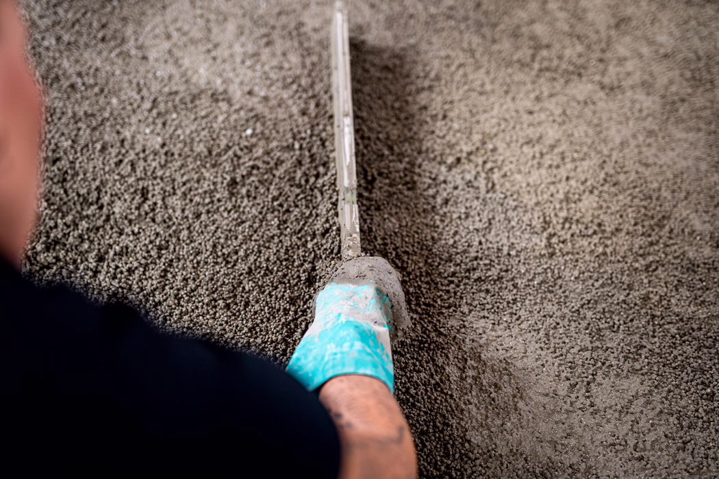 UZIN_Application Image_Screed_Turbolight-System_2023_02 (93) Close-up top-down view of a construction worker's foot wearing a turquoise protective shoe, using a long straight metal screed to level freshly poured wet concrete or levelling compound; the coarse, damp granular texture fills the frame while the screed creates a smooth channel through the material, with shallow depth of field focusing on the central tool and surface.