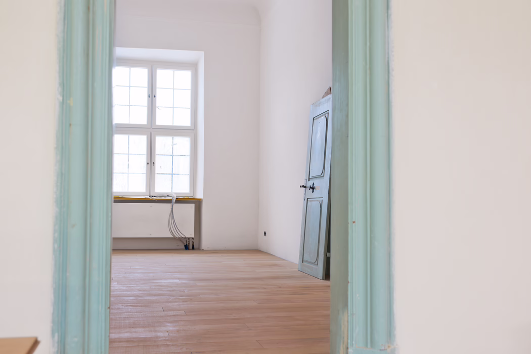 UZIN_Application_Image_MK 200_02_2018-10 Interior view through a pale turquoise painted doorway into an empty, sunlit room. A large four-pane window centered on the far wall fills the space with soft natural light. The room has freshly sanded light wooden floorboards, white plaster walls and a detached, slightly weathered blue-gray wooden door leaning against the right wall. Near the window low heating pipes and a bundle of exposed electrical cables rest on the windowsill and descend to the floor, suggesting ongoing renovation work. The overall atmosphere is calm and minimal, with muted pastel tones and clean architectural lines.