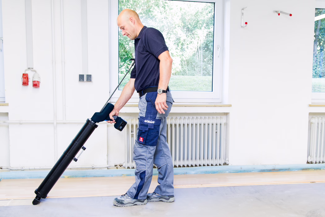 A maintenance worker using a long black floor-cleaning device indoors, standing in front of a window and a radiator, wearing a navy polo, grey work trousers and safety shoes.