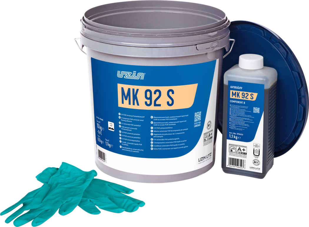 UZIN_Dummy_MK 92 S-10kg_2024-09 Fotografie de produs în studio a unui kit de adeziv pentru parchet: un găleată mare din plastic gri cu o etichetă albastră proeminentă pe care scrie 'MK 92 S', în dreapta o sticlă dreptunghiulară mai mică etichetată 'MK 92 S Component B' cu capac alb, și un capac albastru închis sprijinit în spate. În prim-plan se află o pereche de mănuși de unică folosință turcoaz. Obiectele sunt așezate pe un fundal alb curat și iluminate uniform, evidențiind detaliile ambalajului și informațiile despre produs pentru instalarea pardoselii.