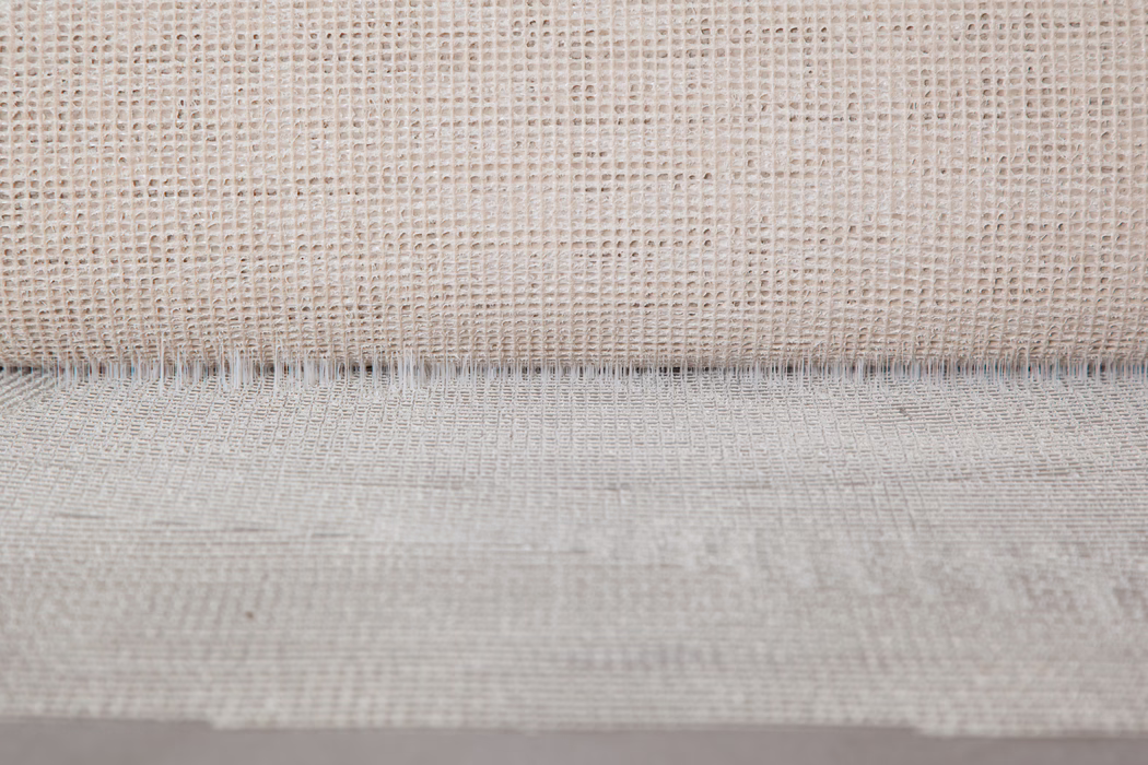 Close-up of a neutral beige woven fabric with an open, grid-like weave. The top shows a rolled edge creating a fine shadow line, while irregular threads and natural fiber variations give a tactile, rustic appearance. Soft, even lighting highlights the texture and subtle depth.