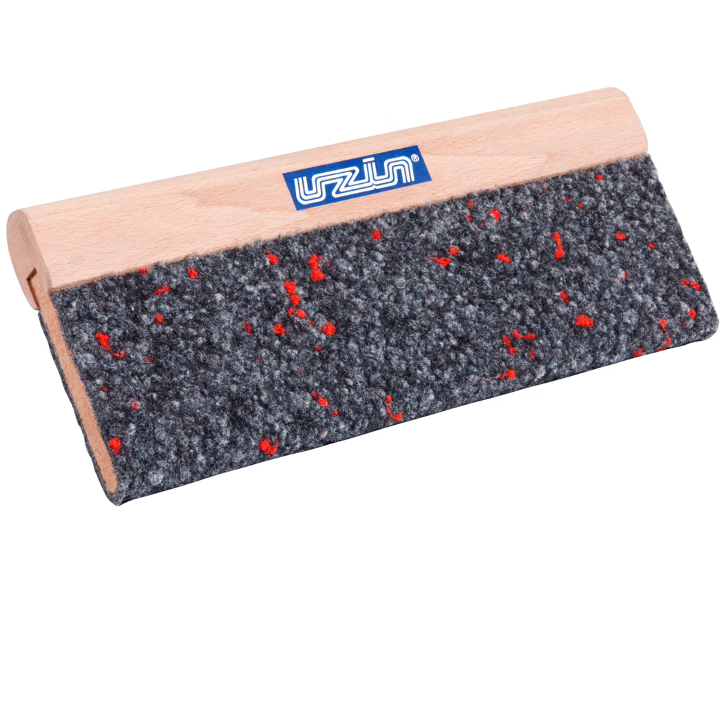 Close-up of a rectangular tool with a light wooden handle and a coarse grey felt pad speckled with small red fibers; a small blue brand label is visible on the smooth beech wood handle. The tool is photographed at an angle on a white background, emphasizing the felt's rough texture and the fine wood grain of the handle.