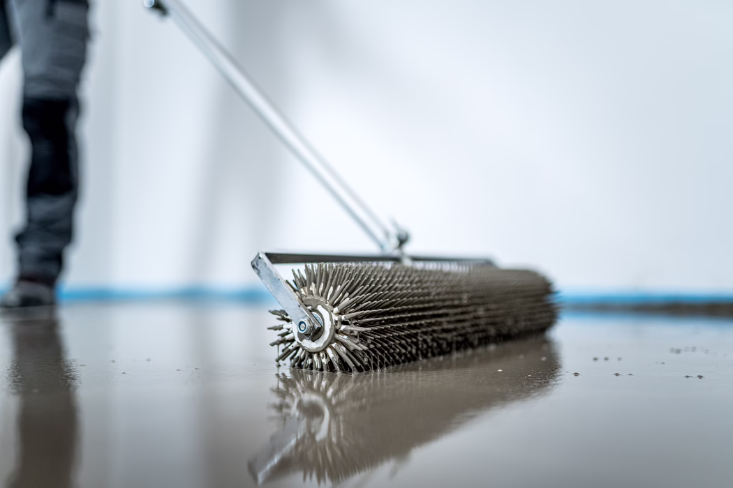 UZIN_Application Image_Screed_Turbolight-System_2023_02 (173) Close-up of a spiked metal roller used to level or texture wet concrete on a floor; the roller rests on a glossy, still-wet surface with a long handle extending out of focus and a blurred worker in the background, soft cool lighting and shallow depth of field emphasize the spikes and their reflection.